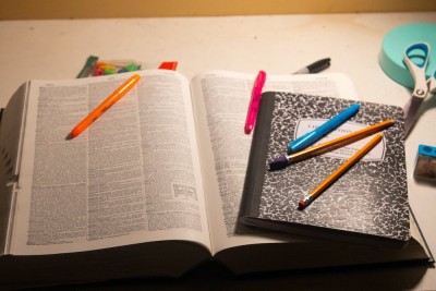 A composition notebook is laid on top of an open dictionary with some pencils and highlighters on top of them both.