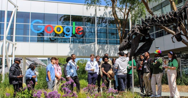 'The Silicon Valley Maymester class stands under a "Google" sign on the Google campus. Their tour guide speaks about a large dinosaur skeleton – a T Rex – that is a focal point of the campus tour.'